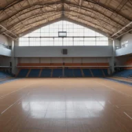 Quadra de ginásio esportivo coberta vazia, com piso de madeira demarcado e arquibancadas ao fundo