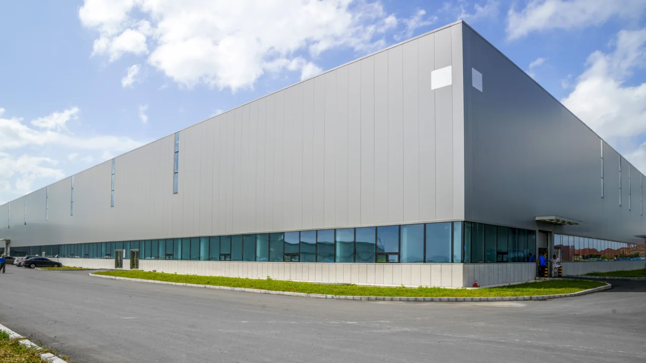 Fachada de galpão industrial moderno sob céu azul, com pátio asfaltado e área verde na entrada