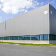 Fachada de galpão industrial moderno sob céu azul, com pátio asfaltado e área verde na entrada