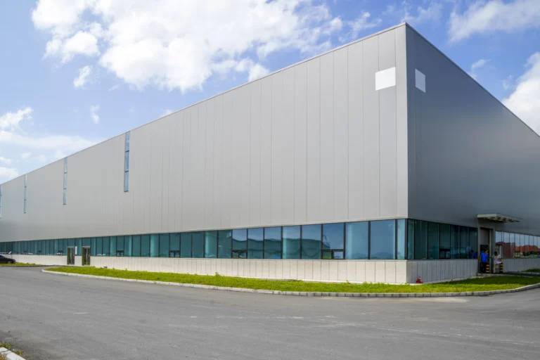 Fachada de galpão industrial moderno sob céu azul, com pátio asfaltado e área verde na entrada